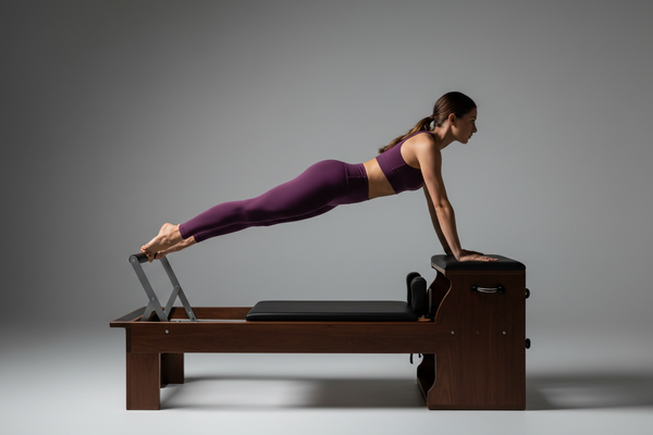 Woman in solid deep plum purple high-compression matching activewear set performing a plank on a wooden Pilates reformer machine in professional studio