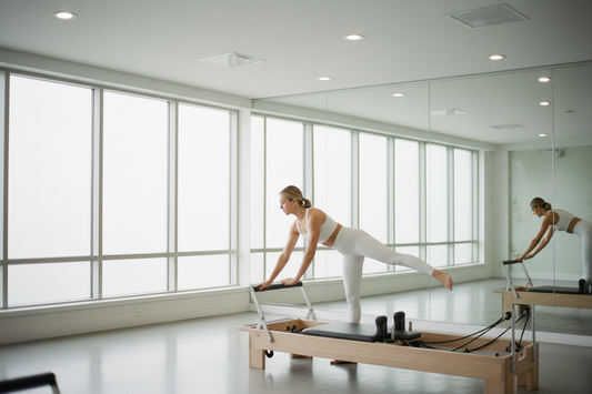 Woman in white matching activewear set performing an elegant leg extension on a wooden Pilates reformer in a bright minimalist studio with floor to ceiling windows | Diva Goddess