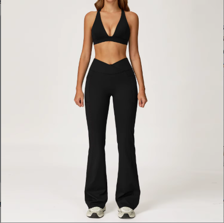 A woman modeling a black two-piece activewear set consisting of a halter-style sports bra and matching high-waisted flared leggings, standing against a plain light gray studio background.